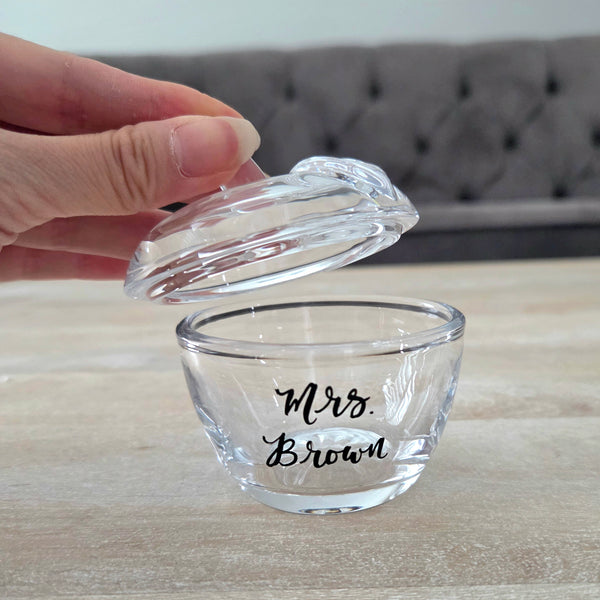 A small clear glass bowl with a matching glass lid, printed with the teacher's name in black script, shown on a light wood surface with a hand lifting the lid.
