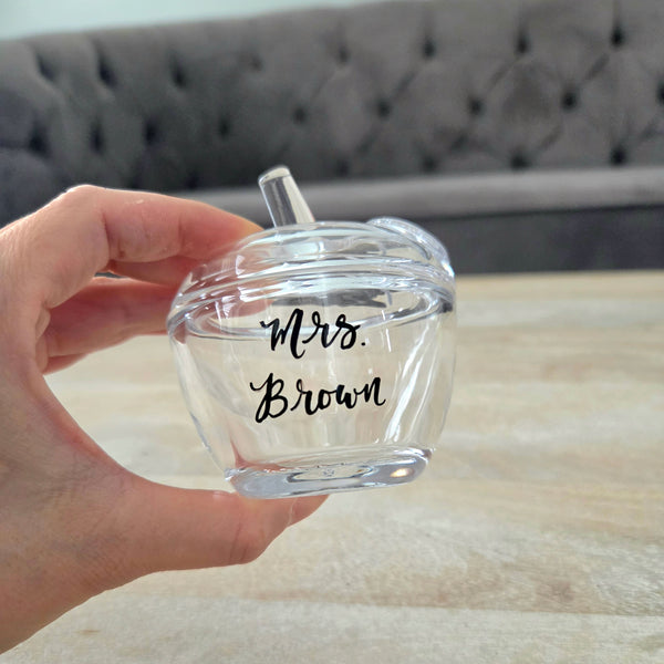A hand holds a small clear glass cup with a lid and straw. The cup is transparent with black script text reading teacher's name