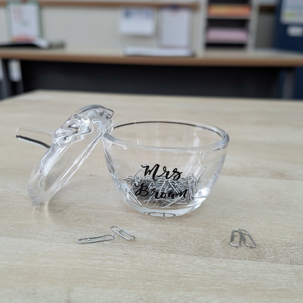 A clear glass jar with its lid leaning against it on a light wood desk, filled with silver paperclips. The bowl is printed with the teacher's name