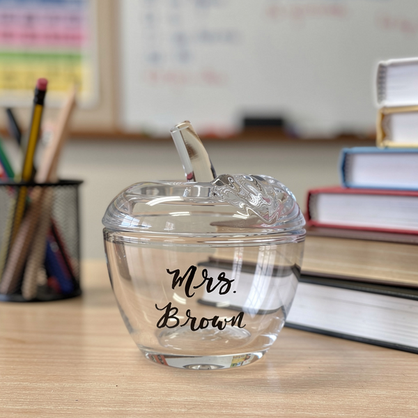 A clear glass apple-shaped bowl with a matching glass lid, featuring teacher's name' in black script on the front, set on a wooden desk with books and a pencil holder in the background.